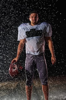 American Football Field: Lonely Athlete Warrior Standing on a Field Holds his Helmet and Ready to Play. Player Preparing to Run, Attack and Score Touchdown. Rainy Night with Dramatic Fog, Blue Light.