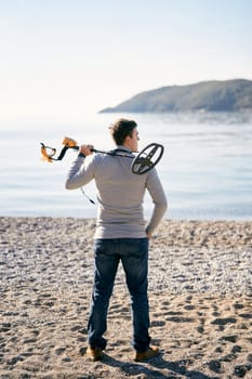 Beach cop with a metal detector on his shoulder stands on the shore and looks at the sea. Back view. High quality photo