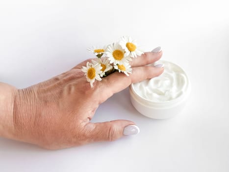Hand middle age woman holding chamomile flowers next to an open jar of white hand cream on white background. Skincare, wellness and beauty product concept. High quality photo