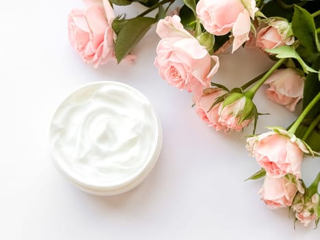 Open jar of white hand, body or face cream next to pink tea roses on white background. Flat lay composition. Skincare and beauty product concept. High quality photo