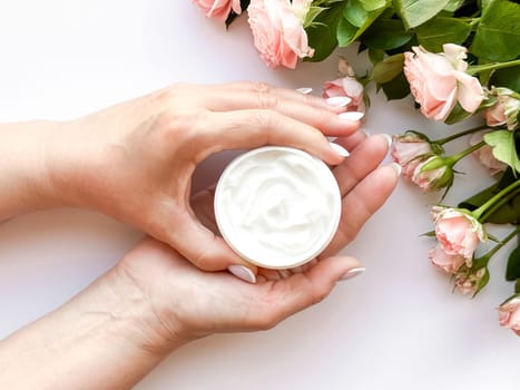 Hands middle age woman hold an open jar of white hand or body cream next to pink tea roses on white background, top view. Flat lay composition. Skincare and beauty product concept. High quality photo