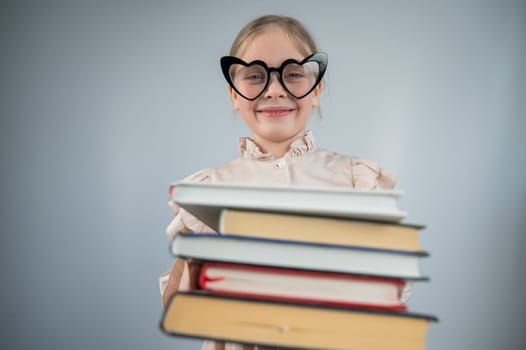 Little girl wearing heart-shaped glasses holding a stack of books on a white background