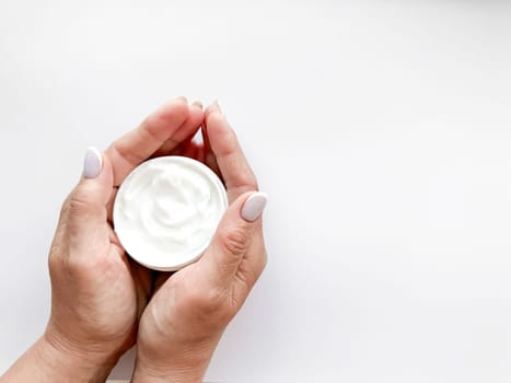 Hands middle age woman hold an open jar of white hand or body cream on white background with copy space. Flat lay composition. Skincare and beauty product concept. Top view. High quality photo