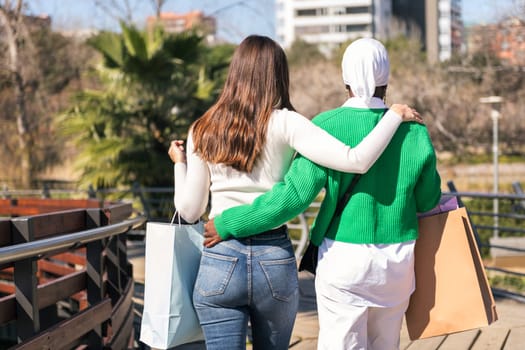 rear view of two unrecognizable young female friends walking through a city park with shopping bags, concept of friendship and urban lifestyle