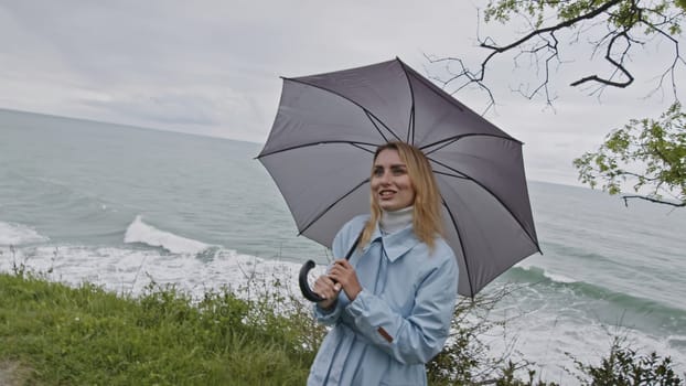 Young smiling woman with eyes full of love spinning grey umbrella. Stock clip. Girl in coat at the ocean coast in spring talking to someone