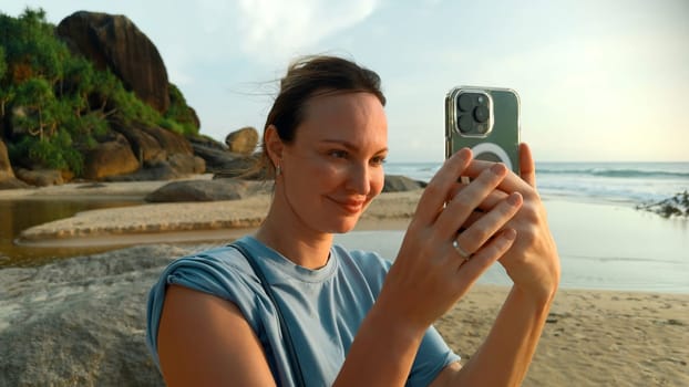 Pretty young caucasian woman takes pictures of the beautiful sea on tropical island beach. Action. Happy girl on vacation photographs on smartphone with palm trees on the background