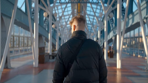 Rear view of man walking in station building. Action. Man walks along sunny corridor of stylish modern building. Beautiful interior of covered bridge of modern city.