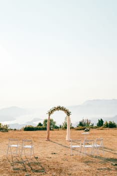 White chairs stand in front of a wedding arch and a reception table with a bouquet of flowers in a clearing in the mountains. High quality photo