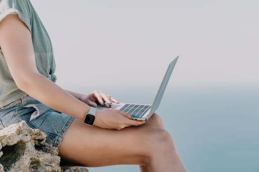 Digital nomad, woman in the hat, a business woman with a laptop sits on the rocks by the sea during sunset, makes a business transaction online from a distance. Freelance, remote work on vacation.