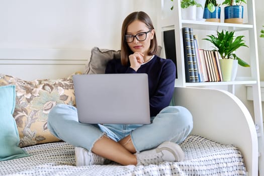 Young woman in glasses working at home, sitting on couch using laptop computer. Freelancing, training, education, remote work, leisure, internet online technology applications apps concept