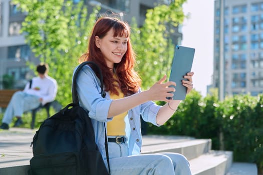 Young attractive female college student using digital tablet outdoor, talking on video call chat conference, educational building background. Education, technology, training, 19,20 years age youth concept