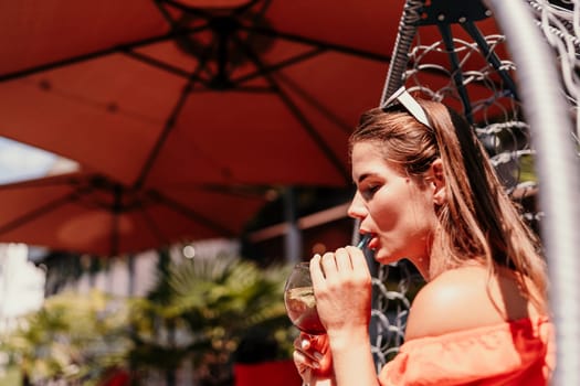 A woman in an orange dress is sitting in a chair with a drink in front of her. She is looking at the camera and she is enjoying her drink. The scene is set in a patio or outdoor area