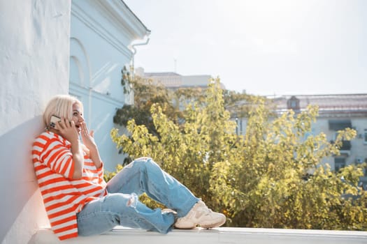 A woman is sitting on a ledge with her phone in her hand. She is wearing a striped shirt and blue jeans. The scene is set in a city with a tree in the background