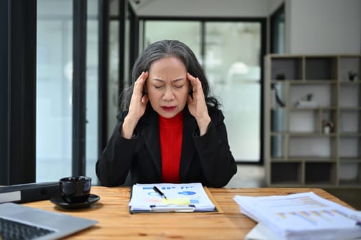Overworked senior businesswoman massaging head feeling stress at workplace.