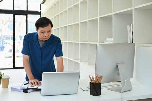 Portrait of senior businessman doing paperwork and using laptop at desk in modern office.