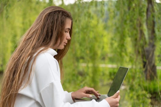 A woman with long hair is sitting on a bench and using a laptop. She is focused on her work, possibly typing or browsing the internet. The scene suggests a sense of productivity and concentration