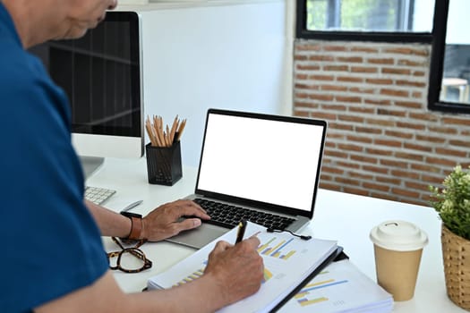 Cropped shot middle age businessman using laptop and analyzing financial reports at desk.