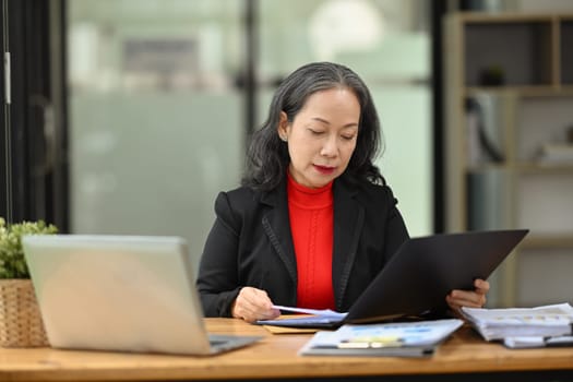 Professional senior woman accounting manager checking financial documents at workplace.