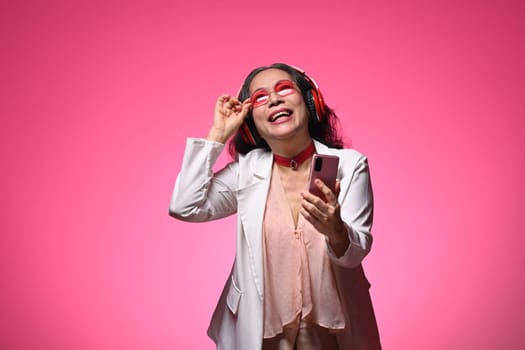 Laughing senior woman in headphone with mobile phone standing against pink background.