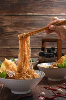 Hand holding spicy egg noodles by chopsticks over wooden rustic background. Asia food and spicy Thai tom yum noodle.