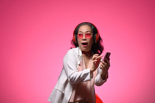 Delighted and surprised senior woman holding smartphone standing against a bright pink background.