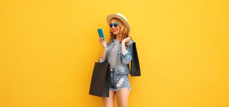 Beautiful happy young woman looking at mobile phone with black shopping bag, wearing denim clothes, summer hat on yellow studio background