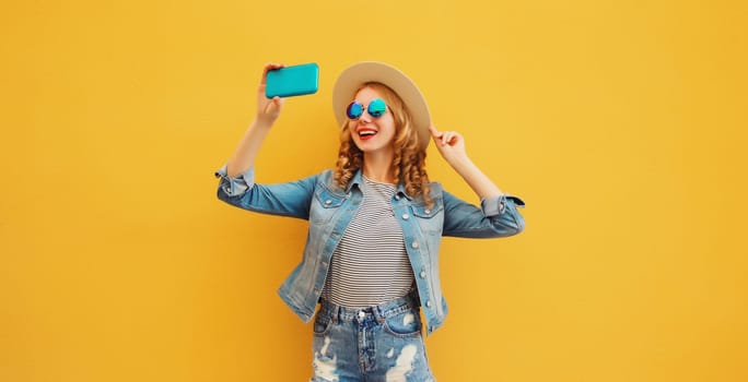 Portrait of happy smiling young woman taking selfie with smartphone wearing summer tourist straw hat, denim clothing on yellow background