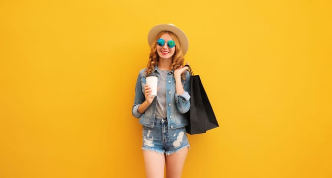 Shopping day! Stylish beautiful happy smiling young woman posing with black shopping bags wearing denim clothing, summer straw hat on yellow studio background
