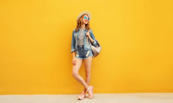 Beautiful young woman full length standing in summer straw hat with backpack, denim clothing, looking away on vivid yellow background