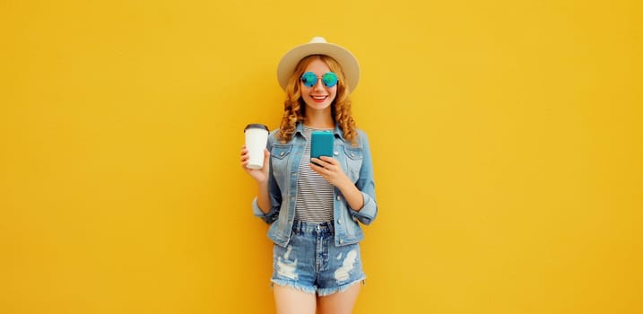 Summer stylish happy young woman with smartphone looking at device holding cup of coffee wearing straw tourist hat, denim clothing posing on yellow background