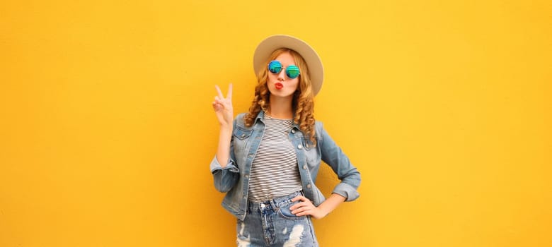 Portrait of beautiful stylish young woman blowing kiss in summer hat, sunglasses, denim jacket posing on yellow studio background