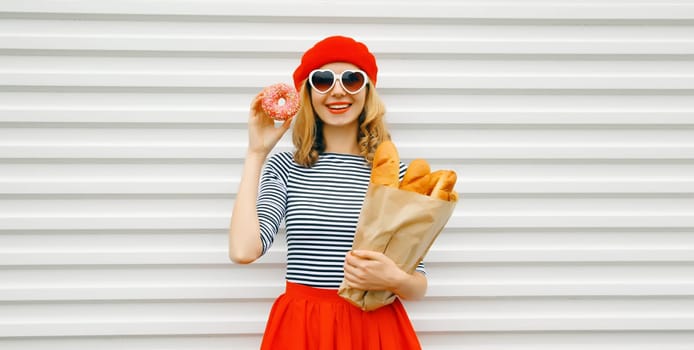 Portrait of happy smiling woman holding grocery shopping paper bag with fresh long white bread baguette eating sweet donut, wear french beret on white background