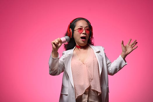 Stylish elderly woman in glasses and headphones singing with microphone over pink background.