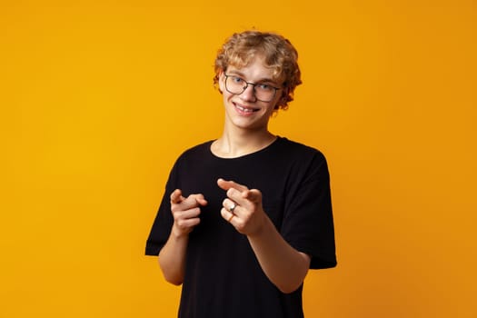 Young curly-haired man in glasses pointing to camera against yellow background close up