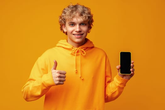 Young smiling man holding smartphone with blank device screen against yellow background close up