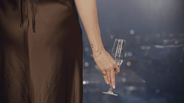 Back view of elegant woman in dress with glass of wine by window. Media. Woman in black dress with champagne in restaurant. Close-up of elegant woman in restaurant on background of window and city.