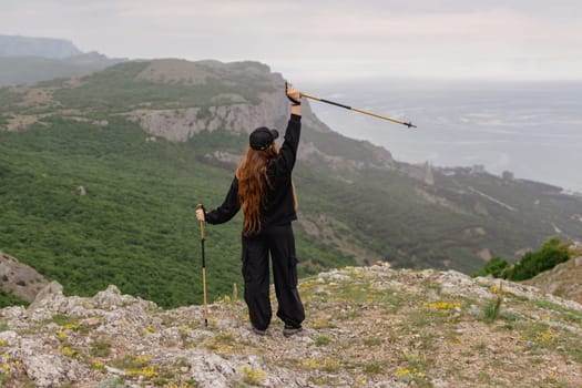 A woman is standing on a mountain top, holding a pair of poles. She is looking out over the ocean, and her arms are raised in the air. Concept of freedom and adventure