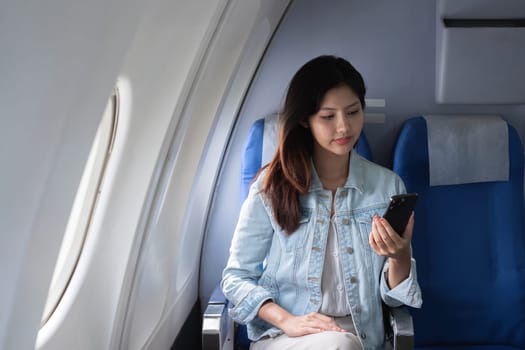 Asian woman using smartphone while sitting in airplane seat. Concept of air travel, technology, and connectivity.