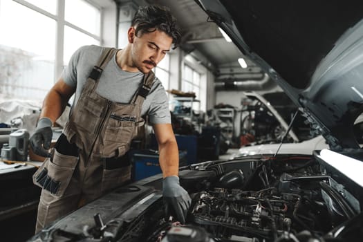 Young male mechanic examining engine under hood of car at the repair garage close up