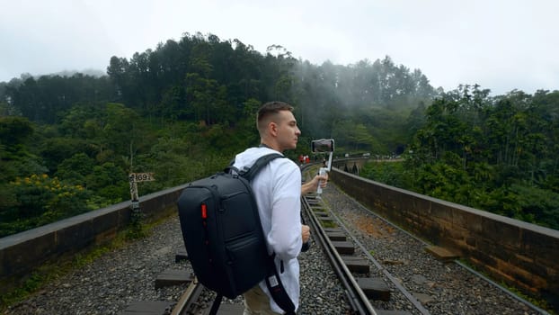 Rear view of a man tourist taking pictures of mountain forested landscape on smartphone. Action. Hiker walking on railways