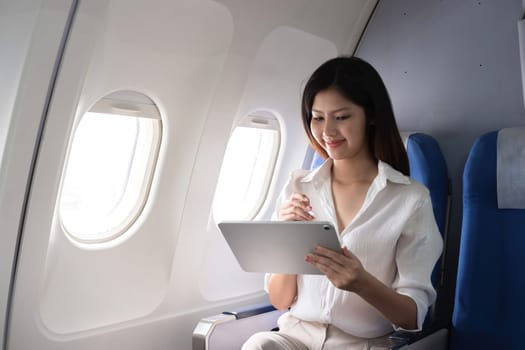 Asian woman using tablet on airplane. Concept of air travel, digital technology, and in-flight entertainment.
