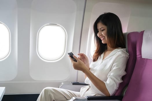 Asian woman using smartphone on airplane seat during flight. Concept of air travel, technology, and connectivity.