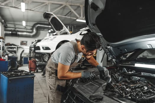 Young male mechanic examining engine under hood of car at the repair garage close up