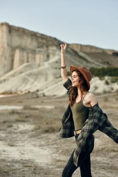 Woman with plaid shirt and hat stands with arms outstretched in desert landscape under clear blue sky