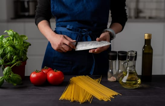 Housewife Checks Knife Sharpness Close Up Near Table With Tomatoes, Pasta, Greens