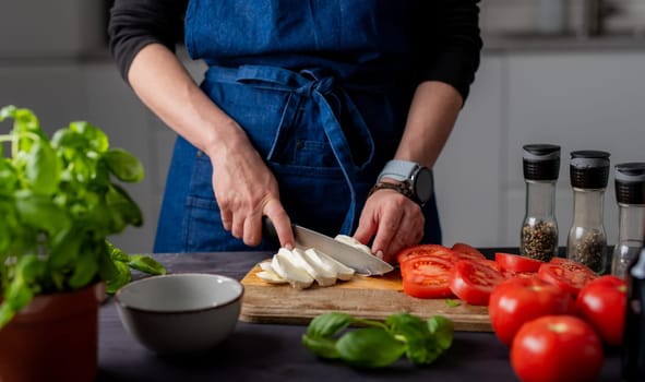 Preparing Caprese Salad, Female Hands Cut Mozzarella Cheese On A Board, With Mozzarella, Basil, And Spices Nearby