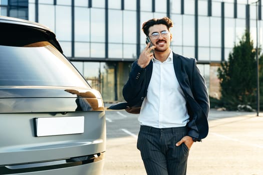 Young businessman in formal suit standing near luxury car and talking on the phone outdoors