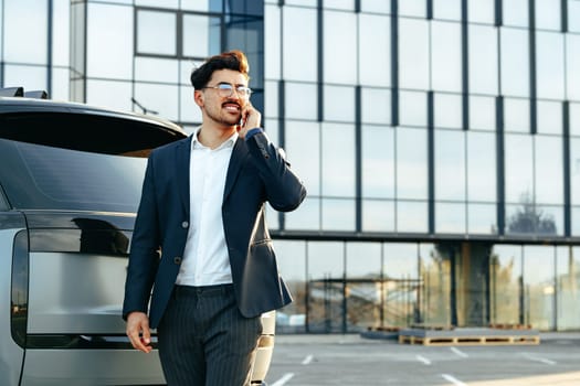 Young businessman in formal suit standing near luxury car and talking on the phone outdoors