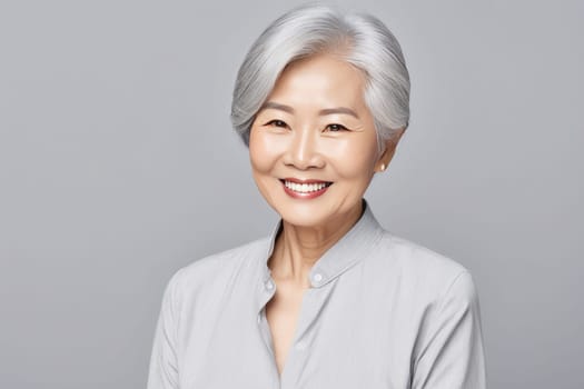 A portrait of an Asian lady, a smiling elderly woman with gray hair, stands on a gray background. A place for text, a banner for advertising