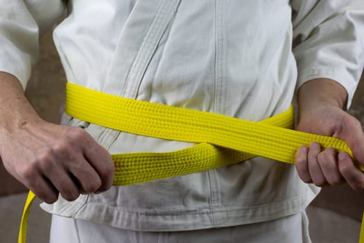 Kyokushinkai karate athlete ties yellow belt around his waist, adjusting martial arts equipment, senior student yellow belt